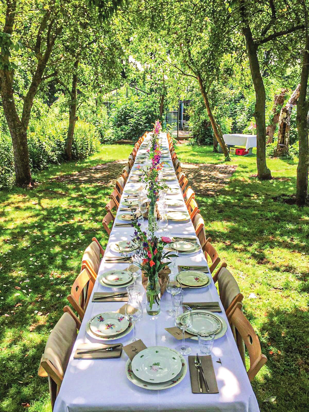Lange tafel met witte tafellakens en veldbloemen in een boomgaard, ingericht door de HALL